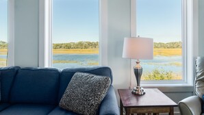 A room with a blue sofa, a patterned pillow, and a lamp on a small table. Large windows provide a view of a natural landscape. - Ocean & Wetland views, 3 decks, steps from the beach, 4 BRs, sleeps 10. (Oak Island)