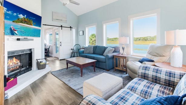 A spacious living room with a fireplace, a large TV, and a view of the outdoors. - Ocean & Wetland views, 3 decks, steps from the beach, 4 BRs, sleeps 10. (Oak Island)
