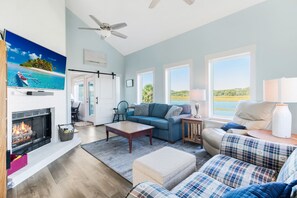 A spacious living room with a fireplace, a large TV, and a view of the outdoors. - Ocean & Wetland views, 3 decks, steps from the beach, 4 BRs, sleeps 10. (Oak Island)