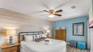 A bedroom with a wooden headboard, a bed with white bedding, a ceiling fan, a dresser, a nightstand, a mirror, and a blue wall. - Tradewinds 14 - Updated Gulf-View Condo, Community Pool, Beach Service Included! (Miramar Beach)