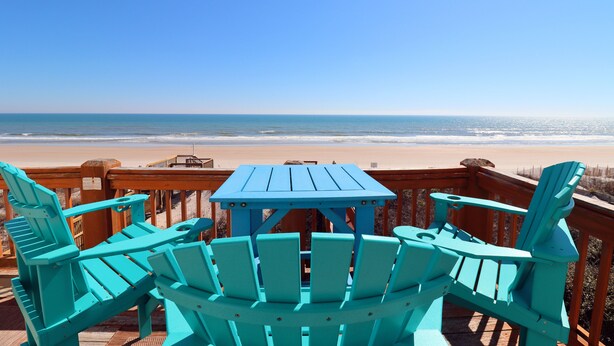 Une terrasse en bois, agrémentée de chaises Adirondack turquoise et d’une table, donnant sur une plage sous un ciel d’un bleu limpide.