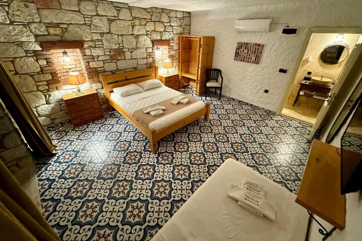 A bedroom with a stone wall, a wooden bed, patterned floor tiles, a wooden nightstand, a chair, and a bathroom visible through an open door.