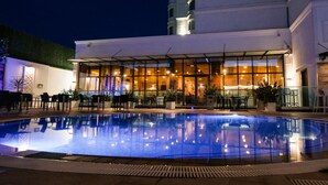 Un espace piscine nocturne doté d’une structure en verre, de sièges extérieurs et d’un bâtiment aux grandes fenêtres en arrière-plan.