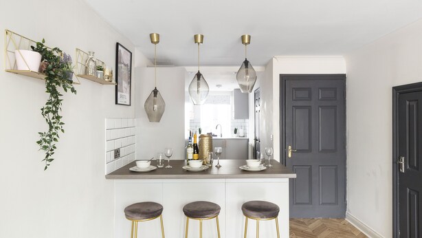 Eine moderne Küche mit weißen Schränken, einem Holzboden im Fischgrätmuster und drei Barhocker mit goldenen Beinen.