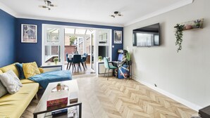 Ein modernes Wohnzimmer mit einem gelben Sofa, einem Fischgrätparkettboden, einem Essbereich mit Tisch und Stühlen sowie einem an der Wand montierten Fernseher.