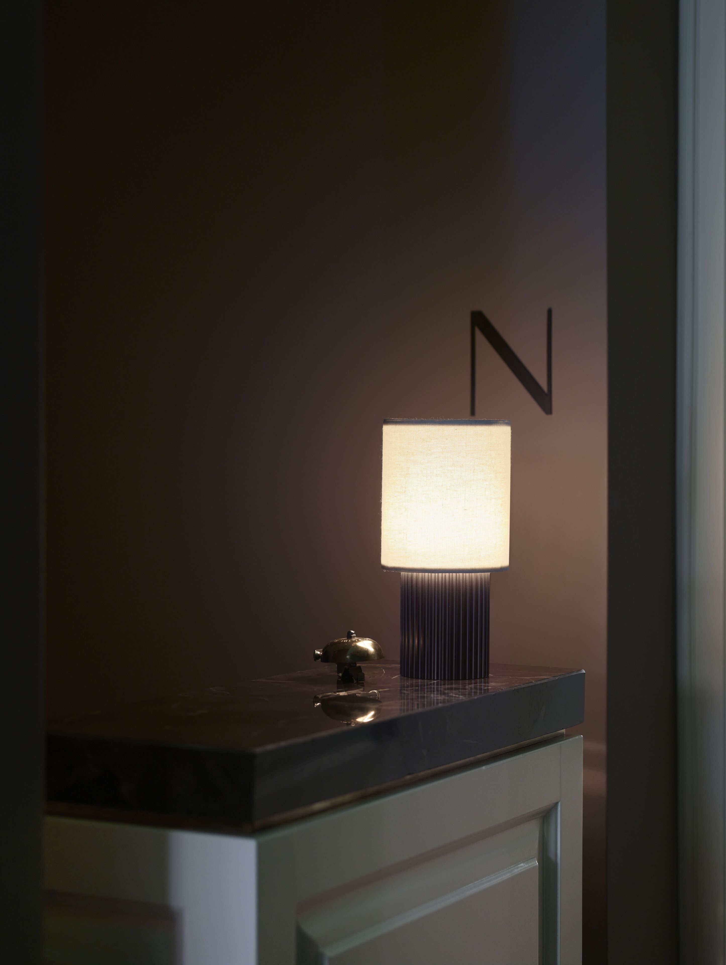 A modern lamp on a dark countertop with a minimalist design, a wall-mounted letter 'N', and a reflective surface below.