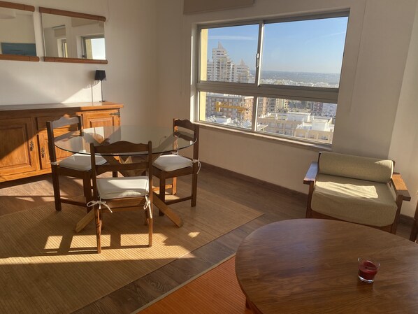 A room with a dining table, chairs, a sofa, a window with a city view, and a wooden cabinet.
