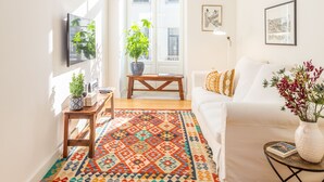 A bright living room with a white sofa, a colorful rug, a wooden coffee table, a potted plant, and a TV mounted on the wall.