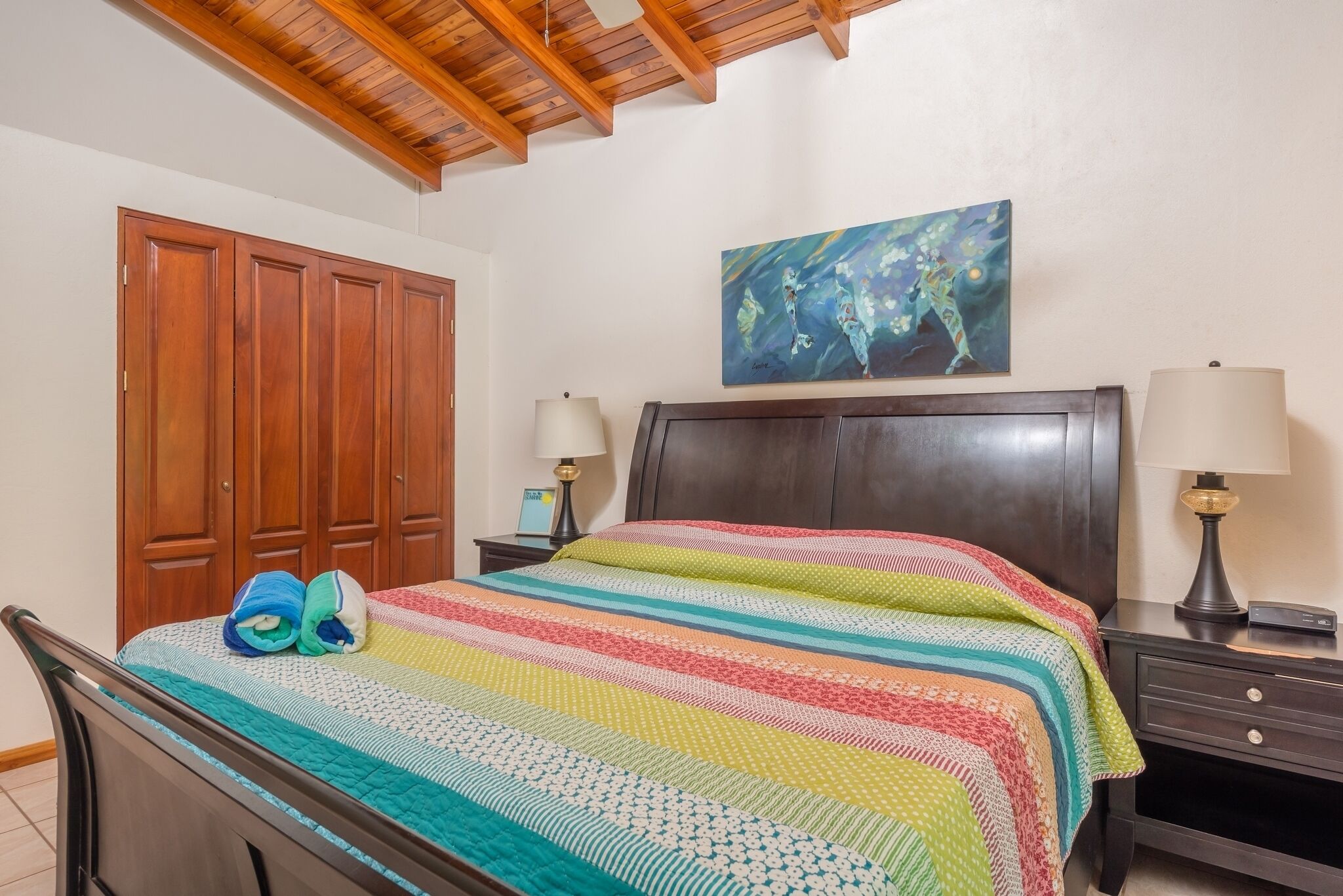 A bedroom with a wooden bed, a colorful striped blanket, a wooden headboard, a nightstand with a lamp, and a painting on the wall.