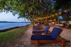 A beachside lounge area with sun loungers and string lights, overlooking a calm sea and distant mountains. - Spacious 1 BR Apartment in Tamarindo, Only Steps from the Beach. (Tamarindo)