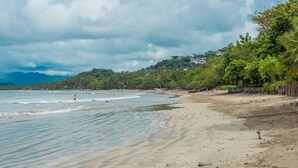Una spiaggia con una battigia sabbiosa, acque calme e colline con edifici sullo sfondo.