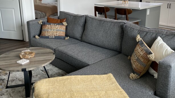 A modern living room with a grey sectional sofa, a round coffee table, and a beige throw blanket. There is a kitchen area with a refrigerator and a dining table. - Oceanside beach retreat 3 min walk to beach Rooftop views (Oceanside)