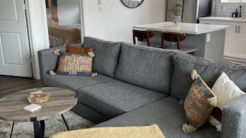 En moderne stue med en grå hjørnesofa, et rundt sofabord og et beige pled. Der er et køkkenområde med et køleskab og et spisebord.