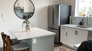 A modern kitchen with a central island, wooden chairs, a refrigerator, and a window with a view. - Oceanside beach retreat 3 min walk to beach Rooftop views (Oceanside)