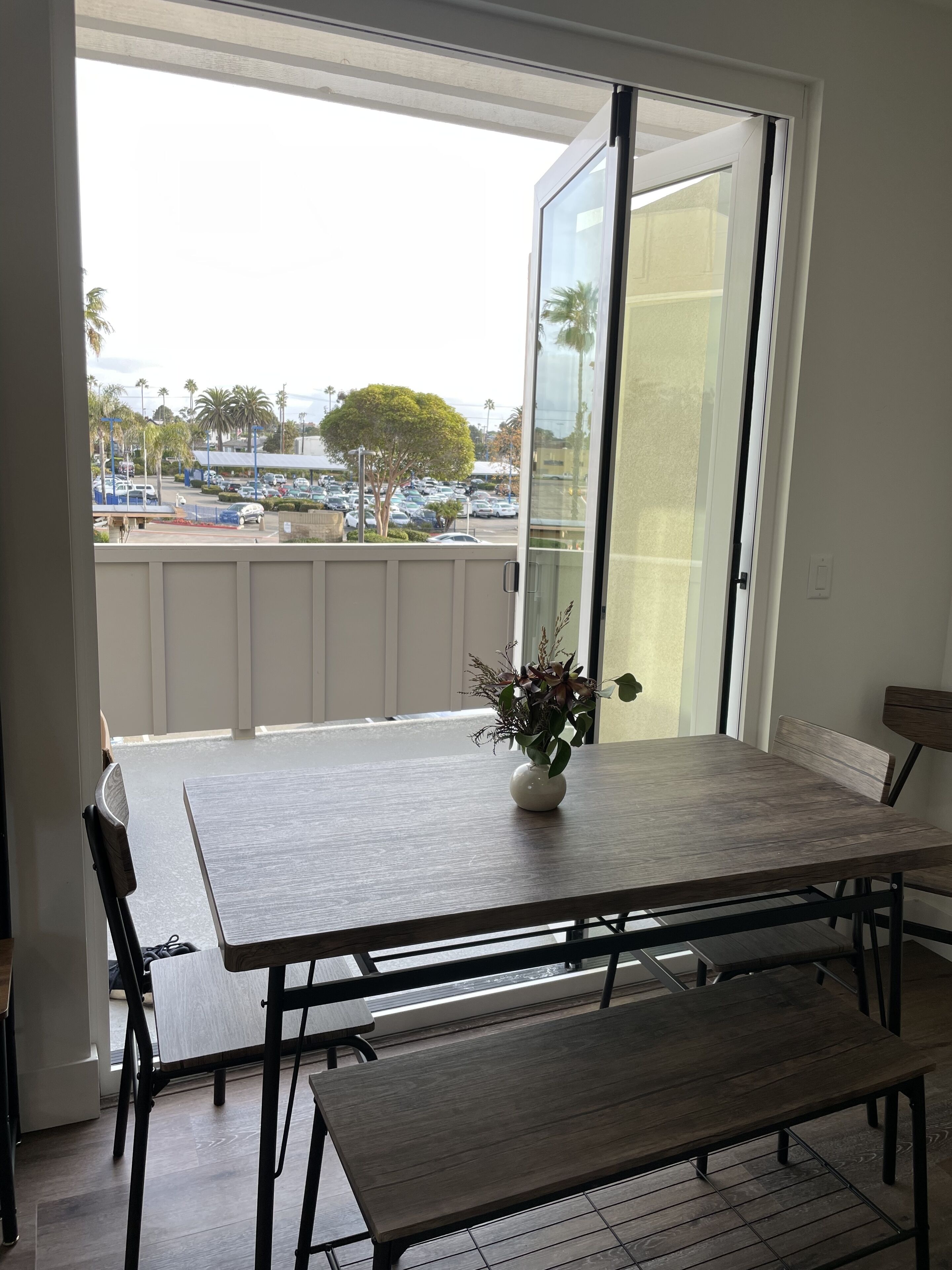 Et spiseområde med et bord og bænke, en vase med blomster og udsigt til en parkeringsplads og træer gennem en skydedør.