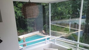 A modern room with a glass wall overlooking a pool and lush greenery. There is a wooden table with a bottle and glasses, a cushioned bench, and a small potted plant. - CARIBBEAN BREEZE CONDO (Akumal)