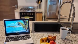 A kitchen counter with a laptop displaying a tropical beach scene, a plate of fruit, and a coffee cup. - CARIBBEAN BREEZE CONDO (Akumal)