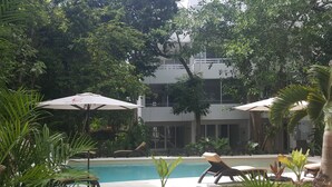 A pool area with lounge chairs and umbrellas, surrounded by lush greenery and a building in the background. - CARIBBEAN BREEZE CONDO (Akumal)