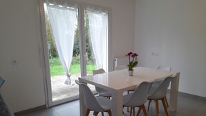Un espace repas moderne avec une table et des chaises blanches, une plante en pot et une porte-fenêtre coulissante donnant sur un jardin.