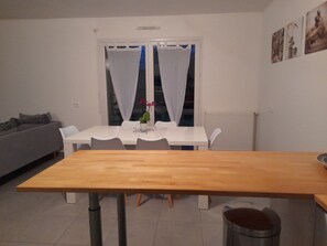 A dining area with a wooden table, white chairs, a small potted plant, and a window with sheer curtains.