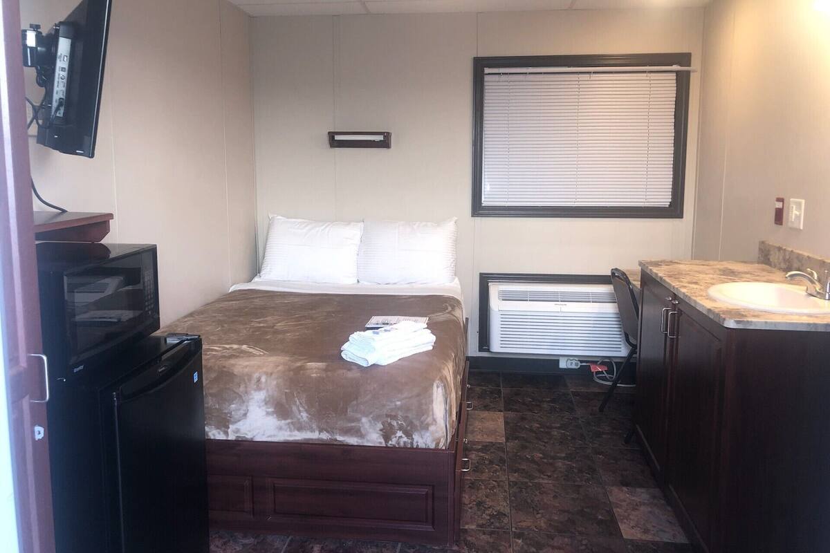 Habitación de hotel pequeña y funcional, con una cama, televisión, microondas, refrigerador pequeño, lavamanos y ventana con persianas.
