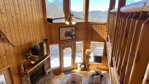 Interior acollidor d'una cabana amb una llar de pedra, un sofà, una televisió i finestrals que ofereixen vistes a l'exterior.