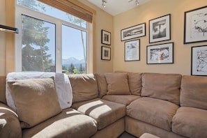 A modern living room with a beige sectional sofa, large windows with a mountain view, and several framed artworks on the walls. - 3 BR 2 BA Ski-in & Ski-out, 50 steps to ski run, unbelievable location! (Mammoth Lakes)