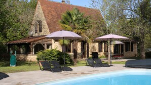 Ein Poolbereich mit Liegestühlen und Sonnenschirmen, ein Steinhaus mit einem Ziegeldach und einer gepflegten Rasenfläche.