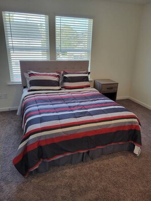 A bedroom with a bed, two windows with blinds, a nightstand, and a carpeted floor. - SC (3745) 2BDR Townhouse Ft Jackson.   (Columbia)
