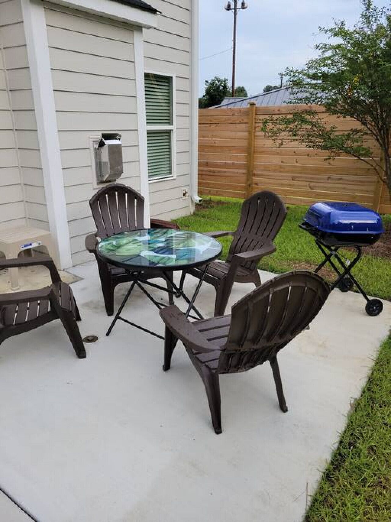 Een terras met een glazen tafel en vier stoelen, een barbecue en een houten omheining op de achtergrond.