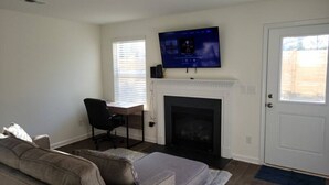 A living room with a sofa, a TV mounted above a fireplace, a desk with a chair, and a door. - SC (3745) 2BDR Townhouse Ft Jackson.   (Columbia)