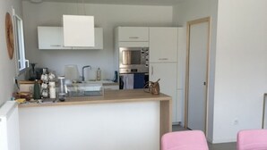 A modern kitchen with white cabinets, a built-in oven, and a countertop with various appliances. There is a dining area with a wooden table and pink chairs. - New house by the sea, Plounéour Brignogan Plages  (Plounéour-Brignogan-plages)
