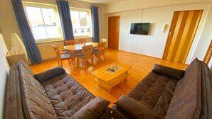 A living room with a sofa set, a coffee table, a dining table with chairs, a TV mounted on the wall, and a window with curtains.