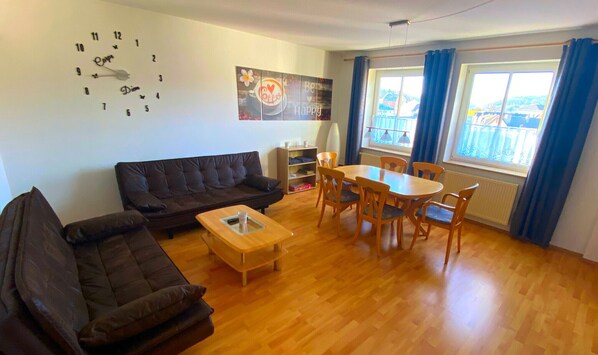 Ein Wohnzimmer mit einem dunkelbraunen Sofa, einem Holztisch, einem Esstisch mit Stühlen, einer Wanduhr und einem Fenster mit Vorhängen.