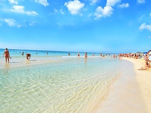 Une plage avec une eau claire et peu profonde, où l’on peut voir des gens se promener et nager, et une rive sablonneuse.