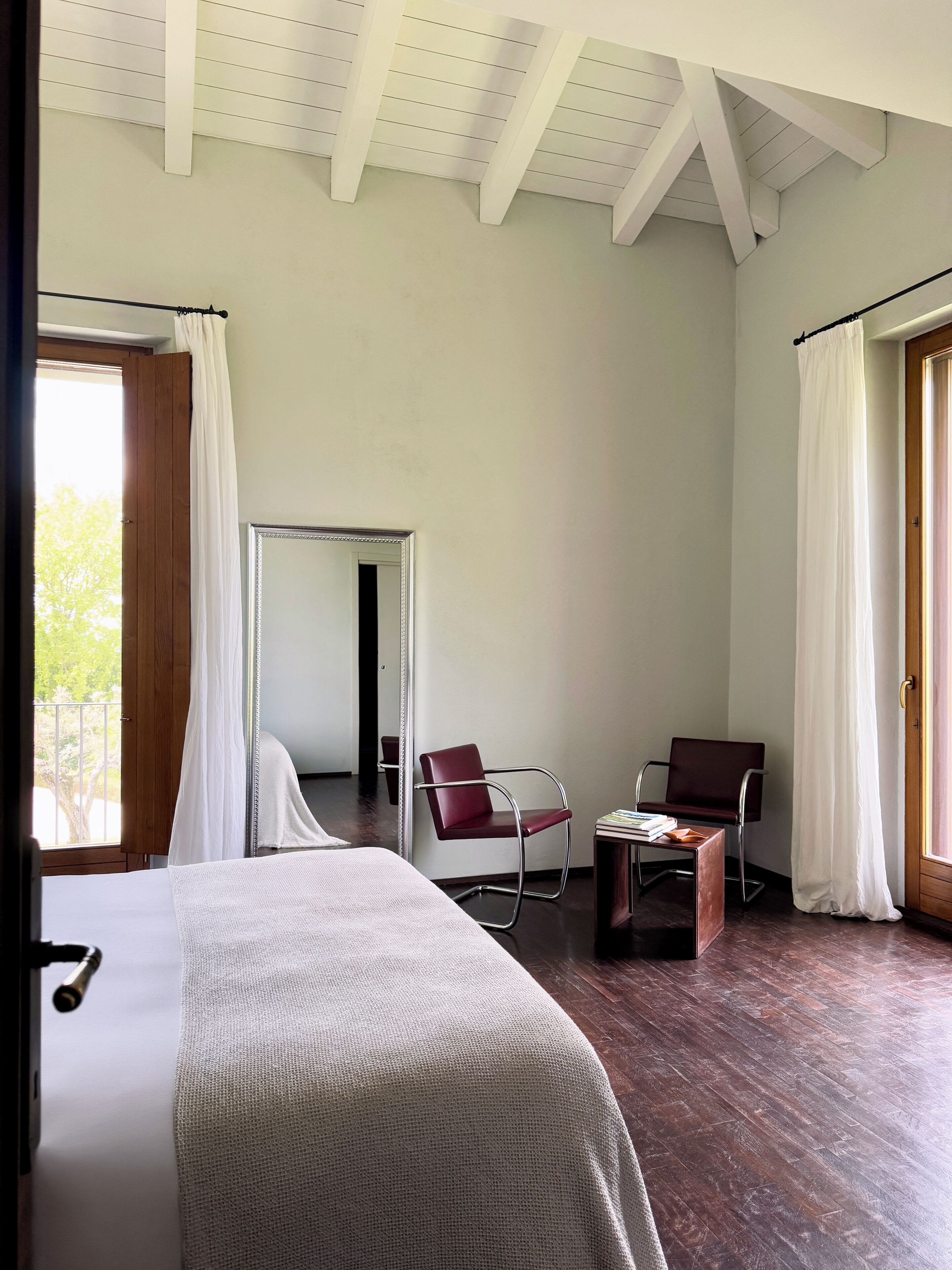 A bedroom with a large bed, a chair, a small table, and a mirror. There is a window with curtains and a door with a wooden frame.