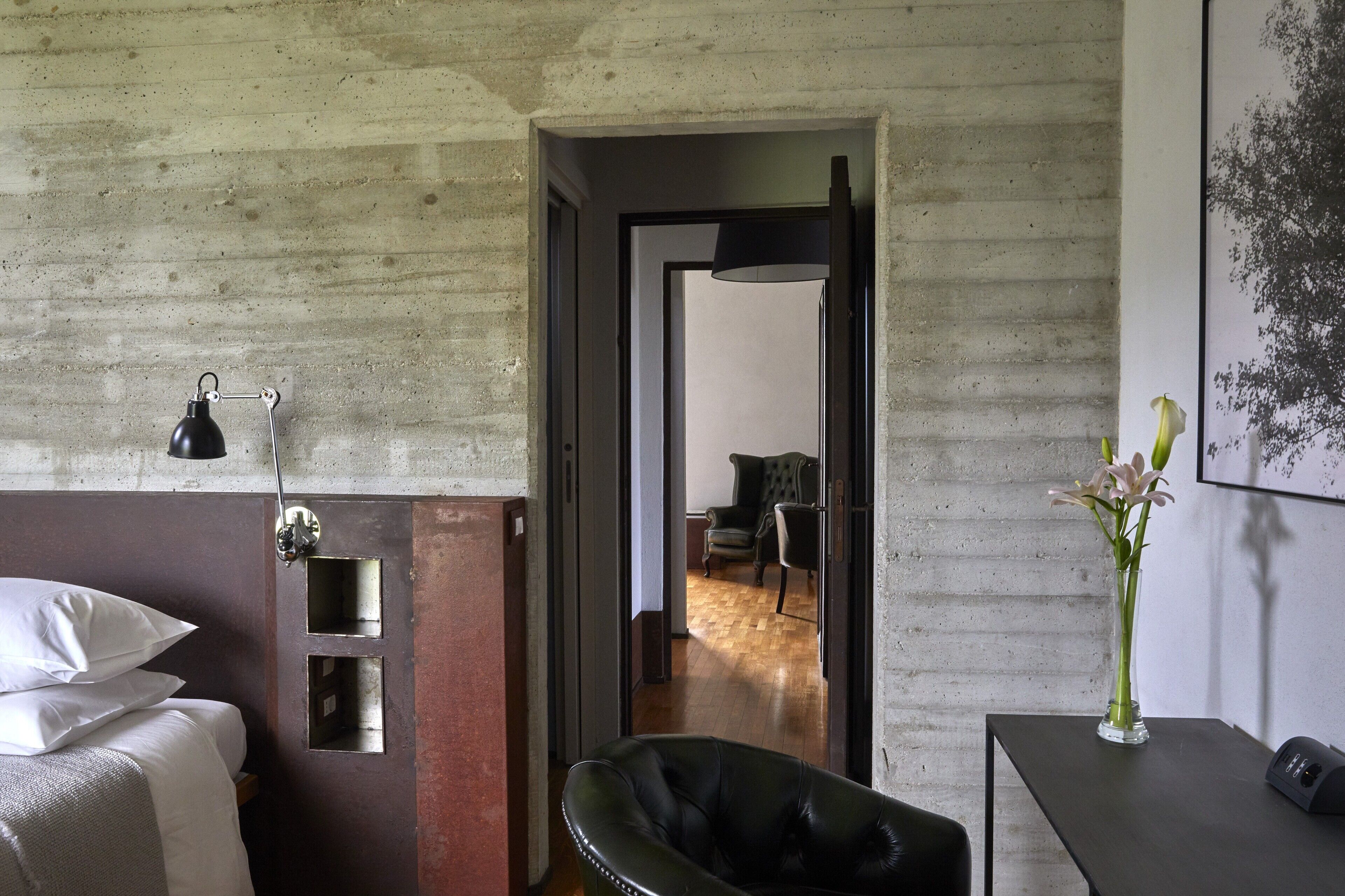 Habitación de hotel con cama, una silla, escritorio con un florero y una puerta que da a otra habitación.