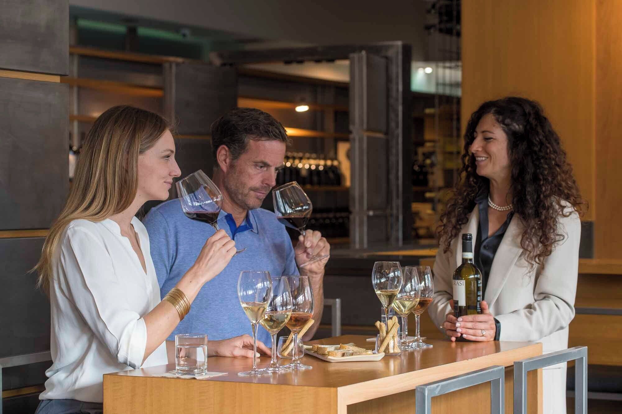 Tres personas en un bar, con copas de vino, una botella de vino y un plato de palitos de pan.