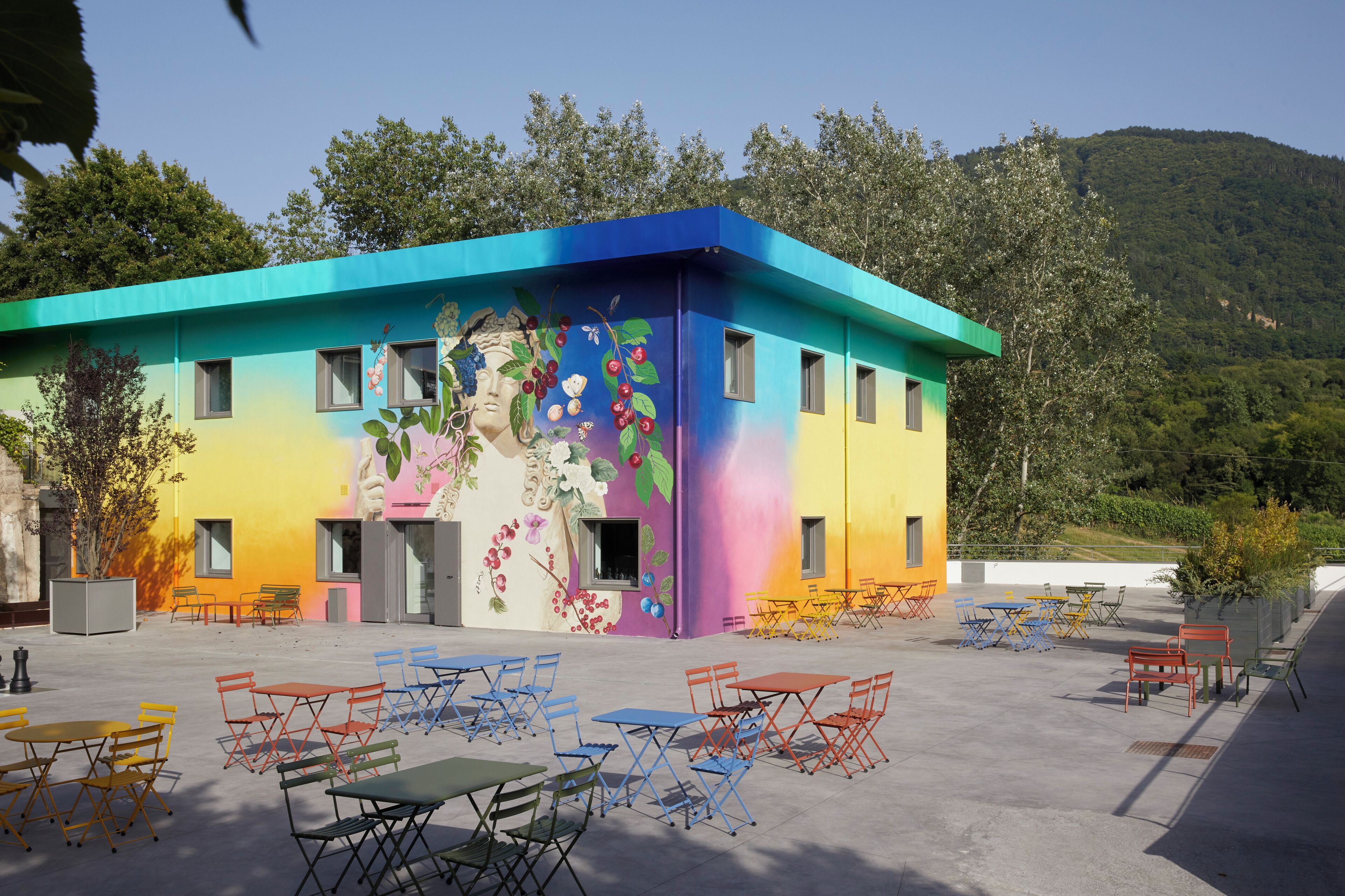 Un bâtiment aux couleurs vives, orné d’une fresque murale, doté d’une terrasse et offrant une vue sur une colline verdoyante.