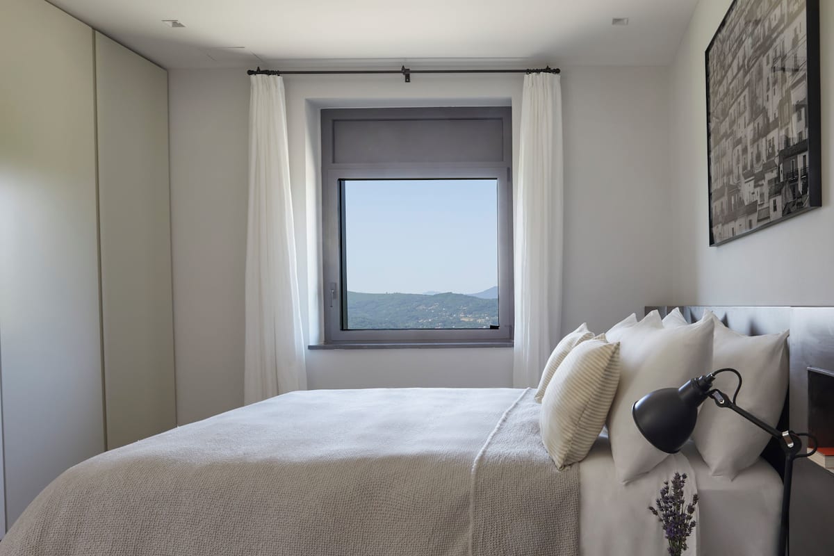 A bedroom with a large window, a bed with white bedding, a black desk lamp, and a framed picture on the wall.