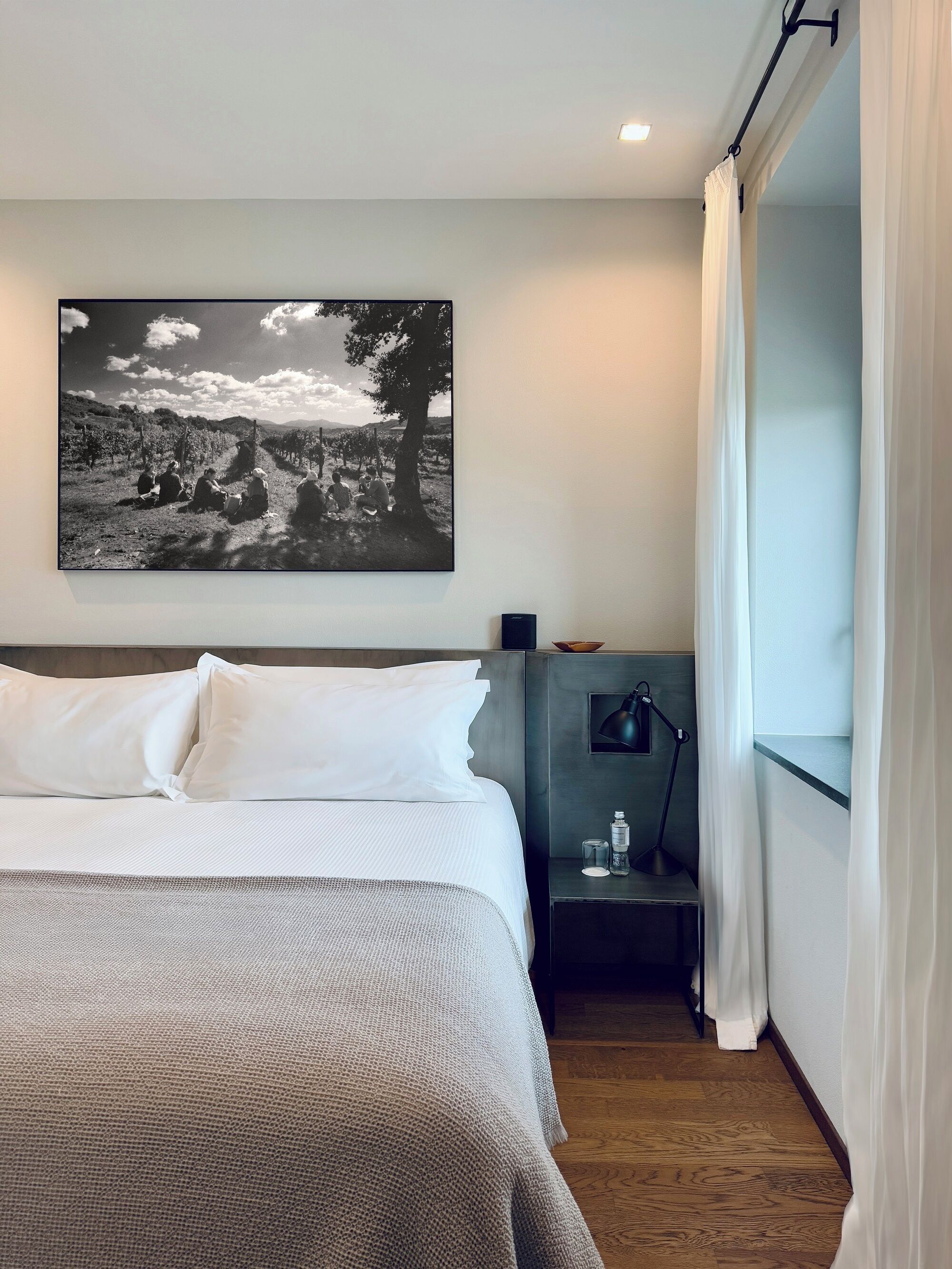 A hotel room with a bed, a nightstand with a lamp, a small table with a bowl, and a large black and white photograph on the wall.