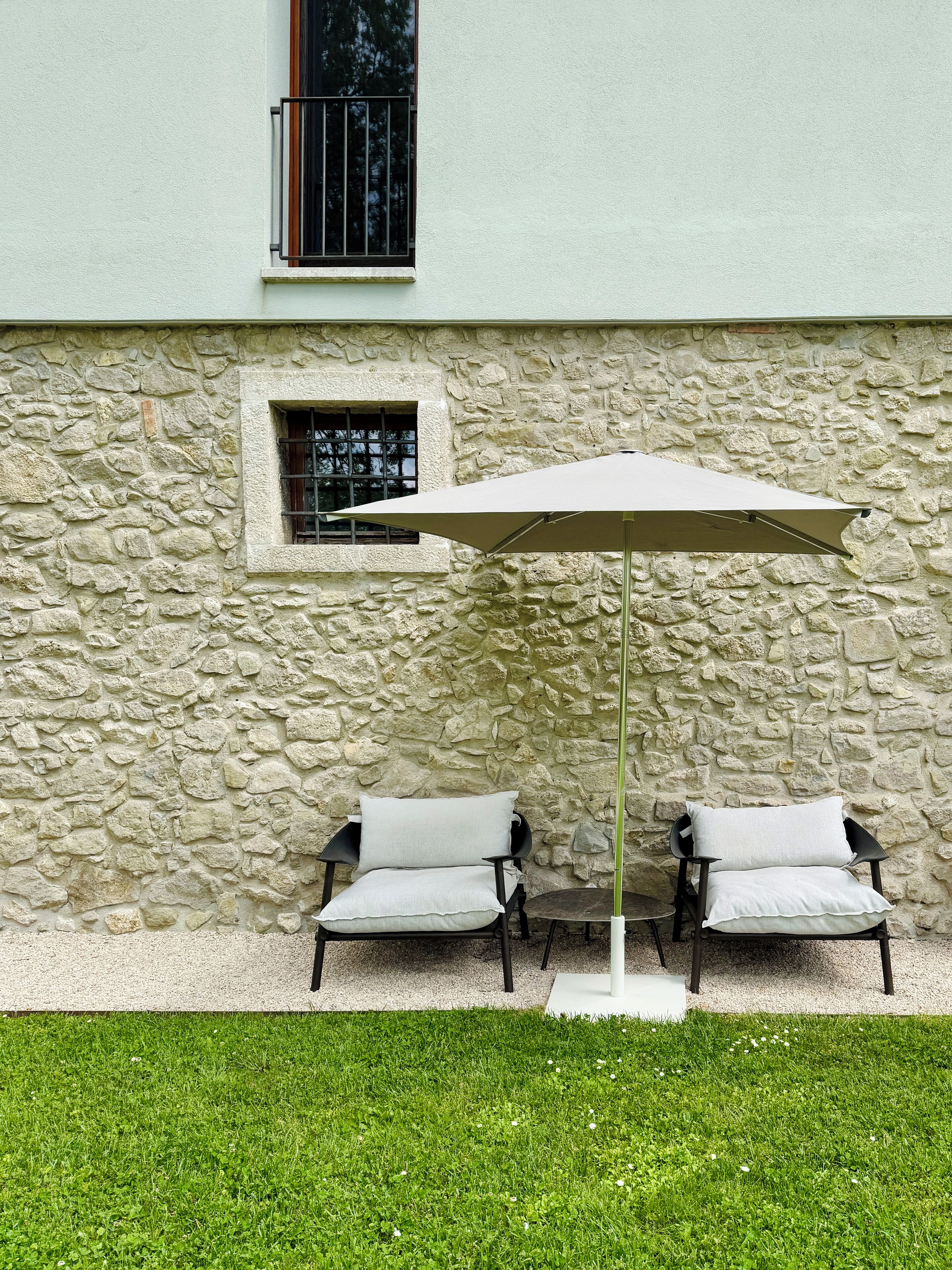 Un edificio de piedra con una ventana y una sombrilla blanca sobre dos sillas.
