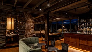 Uma área de bar com balcão de madeira, assentos com mesas redondas e vista para o exterior à noite.