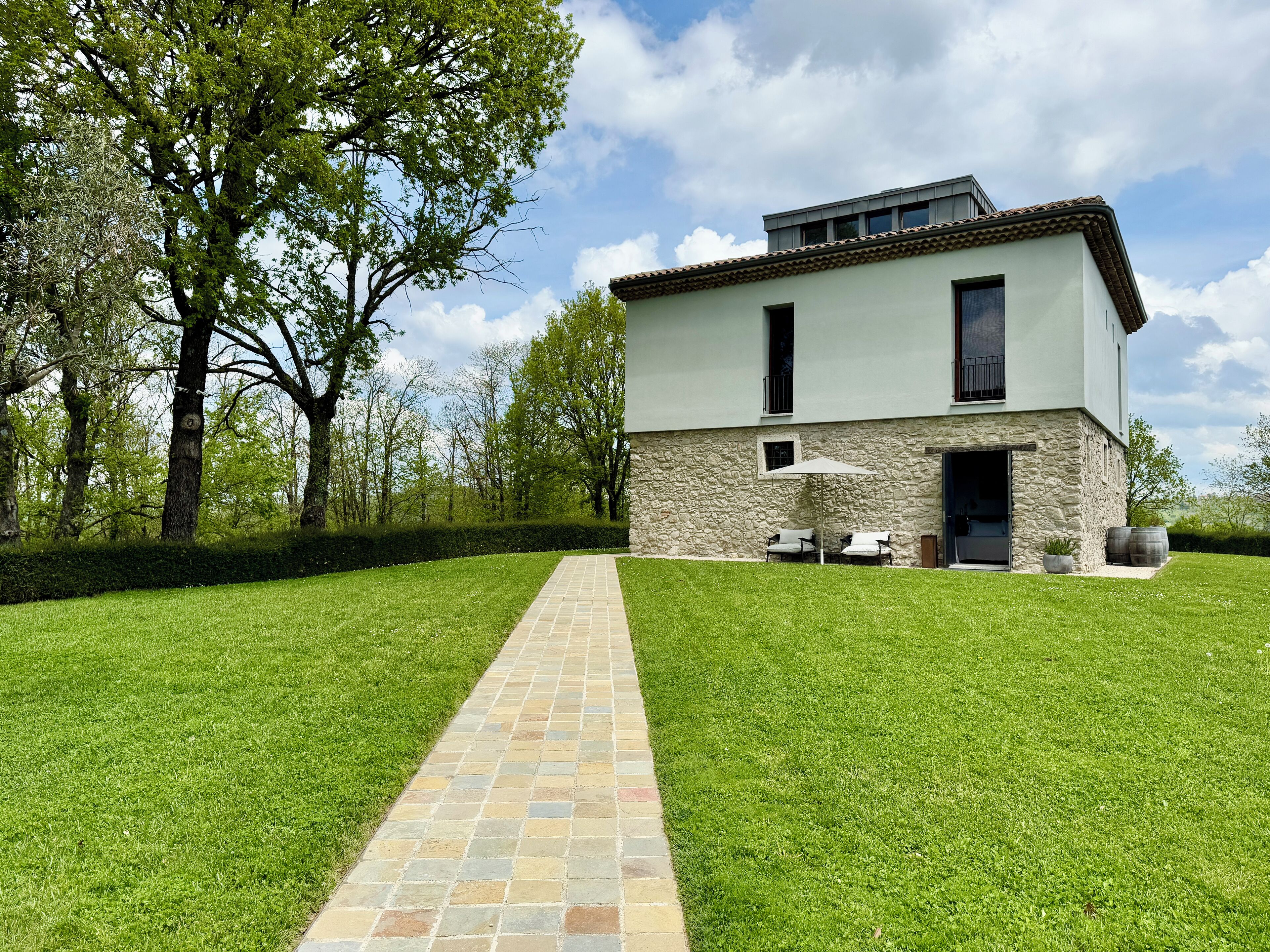 A modern two-story house with a stone facade, a paved pathway leading to a patio with outdoor furniture, and a lush green lawn.