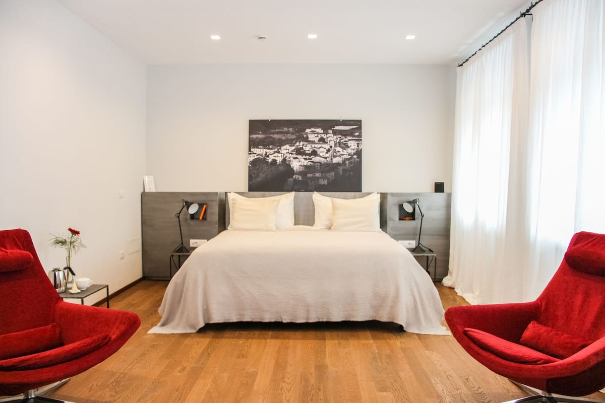 A modern bedroom with a large bed, two red chairs, a nightstand with a lamp, and a wall-mounted artwork.
