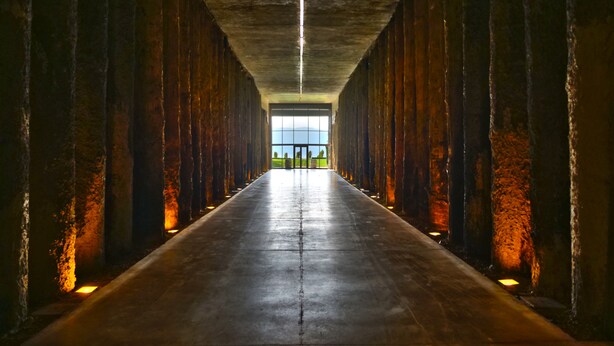 Um corredor comprido e estreito com piso de concreto e paredes, que leva a um espaço amplo e aberto com vista para o verde.