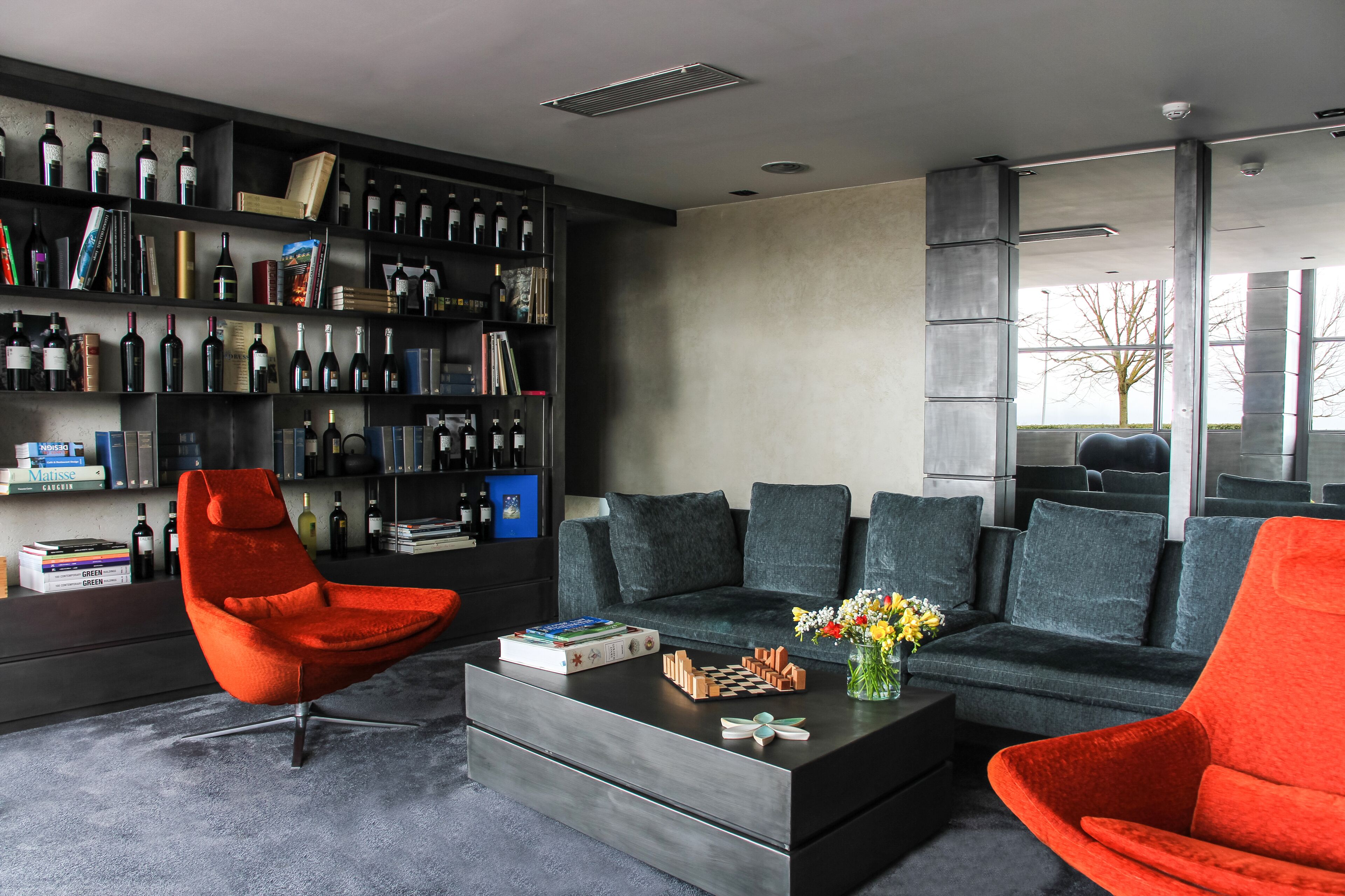 Uma sala de estar moderna com um sofá escuro, uma poltrona vermelha, uma mesa de centro com um jogo de xadrez e uma estante repleta de livros e garrafas de vinho.