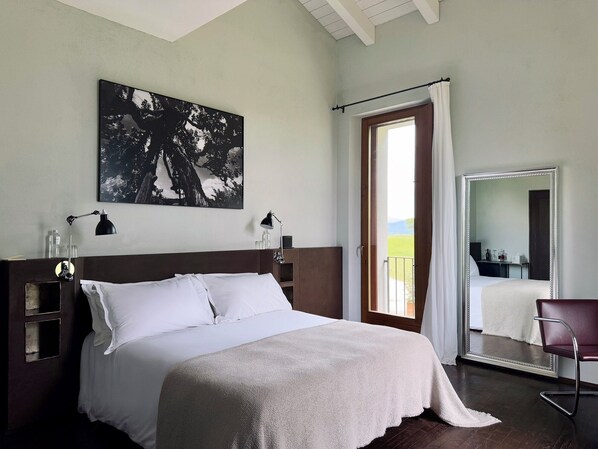 A bedroom with a large bed, a nightstand, a desk, a chair, a mirror, and a window with a view of the outdoors.