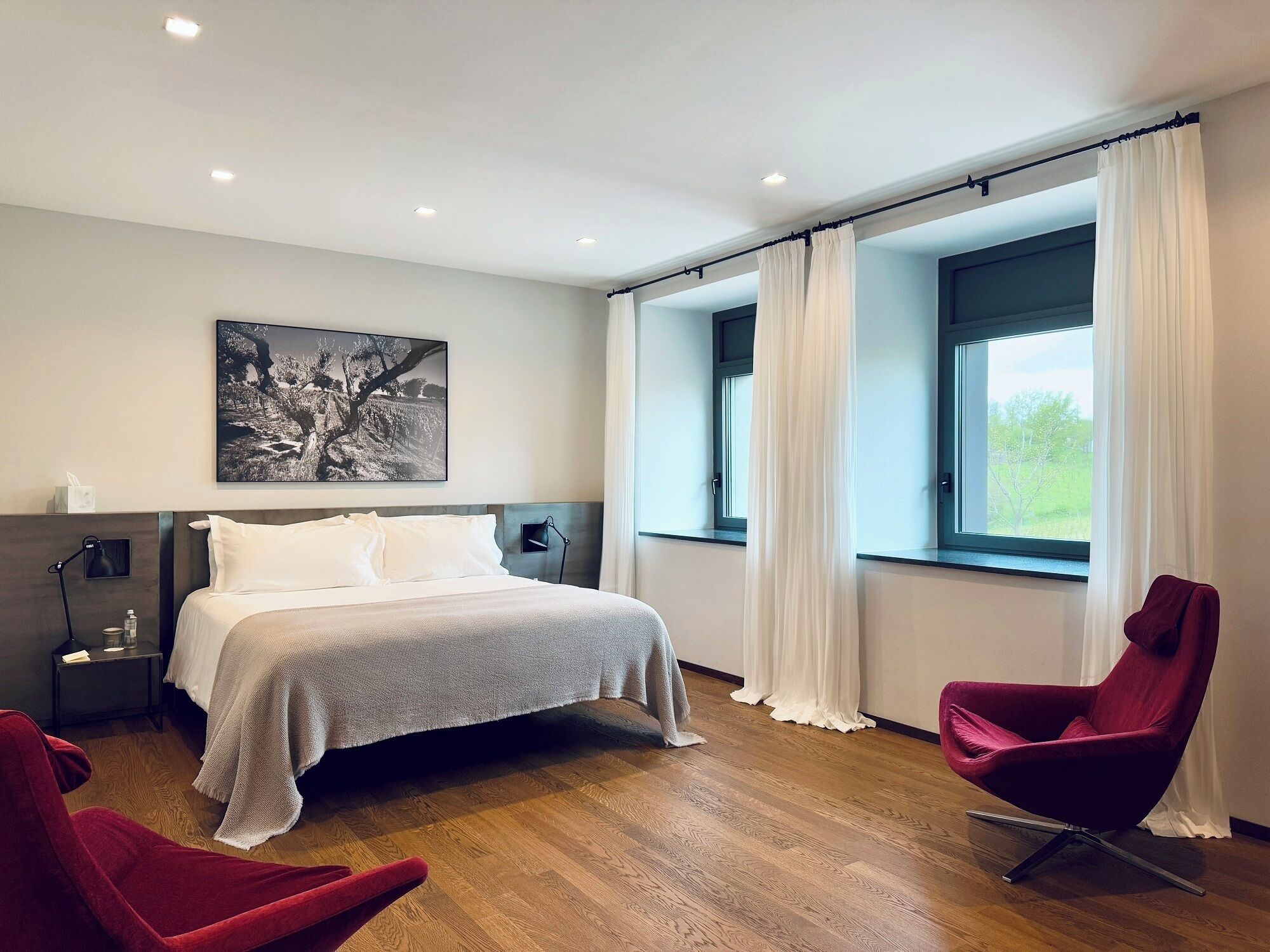 A modern bedroom with a large bed, a nightstand, a red chair, and a window with curtains.