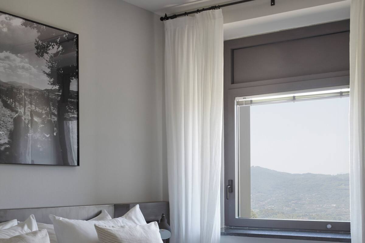 A bedroom with a large bed, white curtains, a window with a view of the landscape, and a painting on the wall.